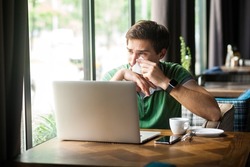 Portrait of upset businessman wearing green T-shirt, sitting in front laptop, crying and cleaning his tears with tissue, expressing sadness. Indoor shot near big window, cafe background.