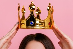 Unknown woman putting on golden crown, arrogance and privileged status, concept of self confidence in success, self-motivation and dreams to be best. Indoor studio shot isolated on pink background.