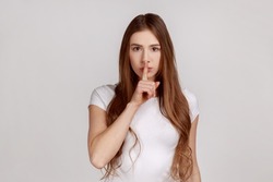Shh, be quiet. Portrait of serious woman showing silence gesture with finger on her mouth, asking to stay quiet, keep secret, wearing white T-shirt. Indoor studio shot isolated on gray background.