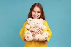 Portrait of little girl hugging cute white toy bear, looking at camera, dreaming, enjoying present, wearing yellow casual style sweater. Indoor studio shot isolated on blue background.