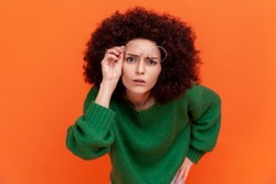 Curious woman with Afro hairstyle wearing green casual style sweater raising her optical glasses, wants to see something better, staring attentively. Indoor studio shot isolated on orange background.