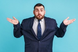 Puzzled clueless man with beard wearing official style suit spreading hands and shrugging shoulders, not sure and indifferent in some questions. Indoor studio shot isolated on blue background.