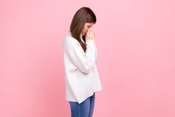 Side view of depressed female crying, felling sorrow, being worried and upset, wipe the tears away, wearing white casual style sweater. Indoor studio shot isolated on pink background.