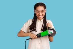 Portrait of smiling woman with dreadlocks holding grinder saw, looking at camera with positive emotions, wearing white shirt and protective glasses. Indoor studio shot isolated on blue background.