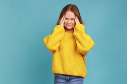 Little girl clutching temples feeling intense headache, child suffering migraine, medical problem, wearing yellow casual style sweater. Indoor studio shot isolated on blue background.