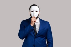 Anonymous unknown businessman covering her face with white mask, hiding her real personality, anonymity, wearing official style suit. Indoor studio shot isolated on gray background.
