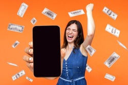 Online investment, winner overjoyed rich woman and money earning. Portrait of amazed young woman showing mobile empty display and celebrating her victory. indoor isolated on orange background.