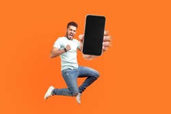 Happy man flying and jumping in air and showing big mobile empty screen for copy space and advertising area. indoor studio shot isolated on orange background