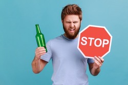 Portrait of bearded man showing alcoholic beverage beer bottle and stop sign, warning and worrying, looking at camera with angry expression. Indoor studio shot isolated on blue background.