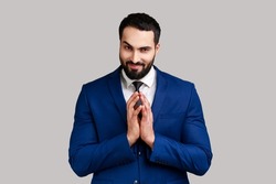 Cunning bearded man clasping hands and planning evil tricky prank or scheming, cheating with sly smile, wearing official style suit. Indoor studio shot isolated on gray background.