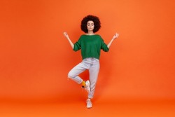 Full length portrait of woman with Afro hairstyle wearing green sweater with mudra gesture hands up, closed eyes, meditating standing in yoga position. Indoor studio shot isolated on orange background
