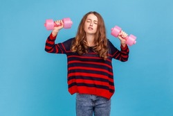 Exhausted woman wearing striped casual style sweater, raising heavy dumbbells with tired expression, does not want to do sport exercises. Indoor studio shot isolated on blue background.