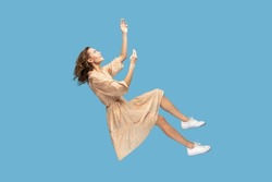 Hovering in air. Relaxed beautiful girl in yellow dress and curly soaring hair levitating, flying in dream with hands up, reaching for something high. indoor studio shot isolated on blue background