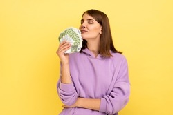 Portrait of attractive young rich woman holding big fan of money, smelling euro banknotes with pleasure, wearing purple hoodie. Indoor studio shot isolated on yellow background.