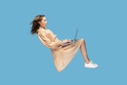 Hovering in air. Surprised girl in yellow dress levitating, looking at laptop screen shocked amazed, surfing web social networks while flying in mid-air. indoor studio shot isolated on blue background