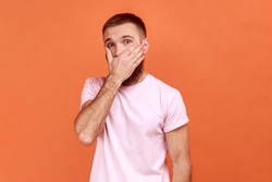 Portrait of bearded man covering mouth with hand to keep silent, afraid to say secret, looking with intimidated expression, wearing pink T-shirt. Indoor studio shot isolated on orange background.