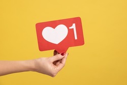 Support interesting web content, push heart button. Closeup of woman hand holding social media like icon, emoji sign to follow subscribe blog. Indoor studio shot isolated on yellow background.