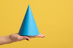 Profile side view closeup of woman hand holding on palm blue birthday cone, celebrating event. Indoor studio shot isolated on yellow background.