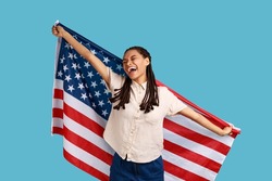 Cheerful attractive woman with black dreadlocks holding USA flag over shoulders and keeps eyes closed and smiling happily, wearing white shirt. Indoor studio shot isolated on blue background.
