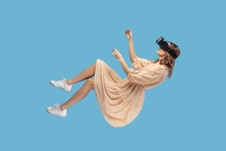 Gamer girl in yellow dress hovering in air, levitating with virtual reality glasses on head, playing game through vr headset, floating in cyberspace. indoor studio shot isolated on blue background
