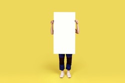 Full length portrait of worker woman covering body with white paper placard with empty space for advertisement or promotional text. Indoor studio shot isolated on yellow background.