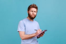 Portrait of bearded man making notes in paper notebook, writing business idea, future plans, looking away with thoughtful expression. Indoor studio shot isolated on blue background.