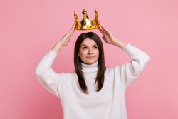 Young brunette woman putting on golden crown, looking with arrogance and smile, privileged status, wearing white casual style sweater. Indoor studio shot isolated on pink background.