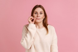 I won't tell anyone. Intimidated blond woman zipping lips and looking at camera, covering mouth promising to keep terrible secret, wearing white sweater. Indoor studio shot isolated on pink background