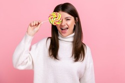 Portrait of brunette female covering eye with big tasty candy, looking at camera, showing tongue out, wearing white casual style sweater. Indoor studio shot isolated on pink background.