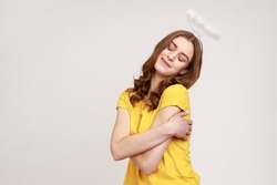 I love myself. Gorgeous angelic teenager girl with halo over head embracing herself lovingly and smiling from pleasure, wearing yellow T-shirt. Indoor studio shot isolated on gray background.