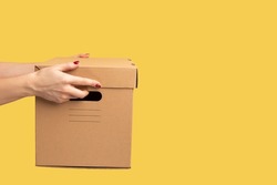 Profile side view closeup of human hand holding cardboard box, carton parcel, delivery concept. Indoor studio shot isolated on yellow background.