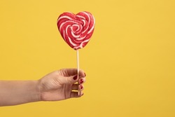 Closeup of woman hand holding appetizing heart shaped candy on stick, big lollipop in arm, confectionery advertising, glucose sweet foods. Indoor studio shot isolated on yellow background.