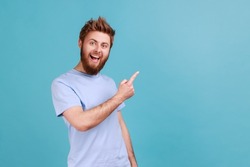 Portrait of positive satisfied bearded man pointing aside, showing blank copy space for idea presentation, commercial text, smiling happily. Indoor studio shot isolated on blue background.
