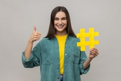 Like and follow trends, popular blog. Optimistic woman holding social media hashtag symbol and thumbs up gesture, wearing casual style jacket. Indoor studio shot isolated on gray background.