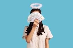 Angelic rich woman with black dreadlocks and nimbus on head hiding behind dollar banknotes, big profit earning cash, wearing white shirt. Indoor studio shot isolated on blue background.
