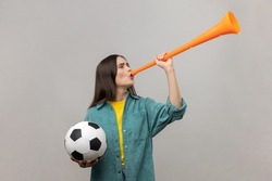 Side view of excited woman blowing in horn holding black and white soccer ball, celebrating victory of favourite football team, wearing casual jacket. Indoor studio shot isolated on gray background.