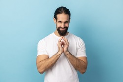 Portrait of devious man with beard wearing white T-shirt smirking and conspiring cunning sly plan, pondering tricky clever idea in mind, wants to cheat. Indoor studio shot isolated on blue background.