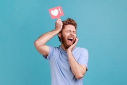 Portrait of excited bearded man holding like counter button, smiling with pleasure, rejoicing and amusing with popularity on internet, social network. Indoor studio shot isolated on blue background.