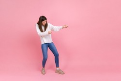 Full length portrait of girl pulling invisible rope with effort, using all strength to achieve goal, wearing white casual style sweater. Indoor studio shot isolated on pink background.