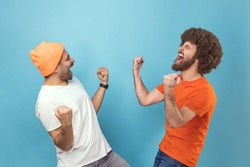 Portrait of two young adult hipster men showing yes gesture and screaming celebrating victory, success, dreams comes true, euphoria. Indoor studio shot isolated on blue background.