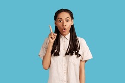 Pleased young woman with black dreadlocks pointing upwards, looking at camera with open mouth, having new great idea, wearing white shirt. Indoor studio shot isolated on blue background.