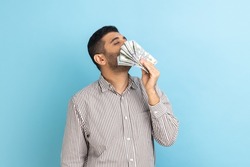 Portrait of handsome greedy arrogant businessman with beard smelling dollar banknotes with expression of pleasure, wearing striped shirt. Indoor studio shot isolated on blue background.