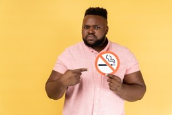 Portrait of serious man wearing pink shirt holding and pointing index finger at no smoking sign in hands, quit smoking, no tobacco day. Indoor studio shot isolated on yellow background.