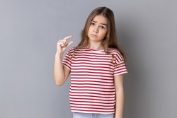 Need some more. Little girl wearing striped T-shirt doing a little bit gesture and looking with displeased imploring expression, showing minimum. Indoor studio shot isolated on gray background.