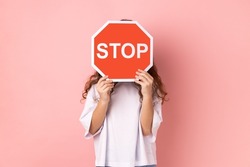 Portrait of anonymous little girl wearing white T-shirt covering face with stop symbol, holding red traffic sign, forbidden access. Indoor studio shot isolated on pink background.