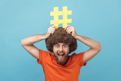 Portrait of man with Afro hairstyle holding yellow hashtag sign board on head, dreaming about target tagging posts on his website, smm manager. Indoor studio shot isolated on blue background.