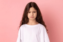 Portrait of upset little girl wearing white T-shirt expressing negative emotions and sorrow, has bad mood, being sad and unhappy. Indoor studio shot isolated on pink background.