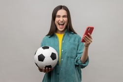 Extremely happy excited woman standing holding soccer ball and using smart phone, betting and winning, wearing casual style jacket. Indoor studio shot isolated on gray background.