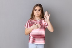 Portrait of serious patriotic little girl wearing striped T-shirt holding hand on heart, swearing to speak truth, honor and conscience. Indoor studio shot isolated on gray background.