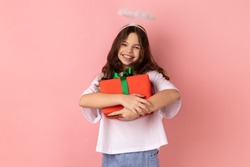 Portrait of little girl wearing white T-shirt and angelic halo embracing wrapped box, enjoying birthday gift, satisfied with long-awaited present. Indoor studio shot isolated on pink background.